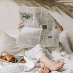 Person relaxing in bed with a newspaper, coffee, and croissant for a calm morning vibe.