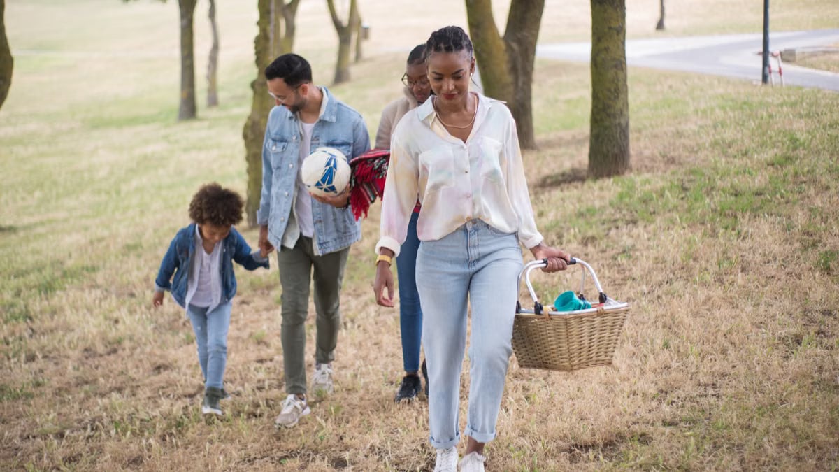 A joyful family walking in a park, carrying a picnic basket and a ball on a sunny day.